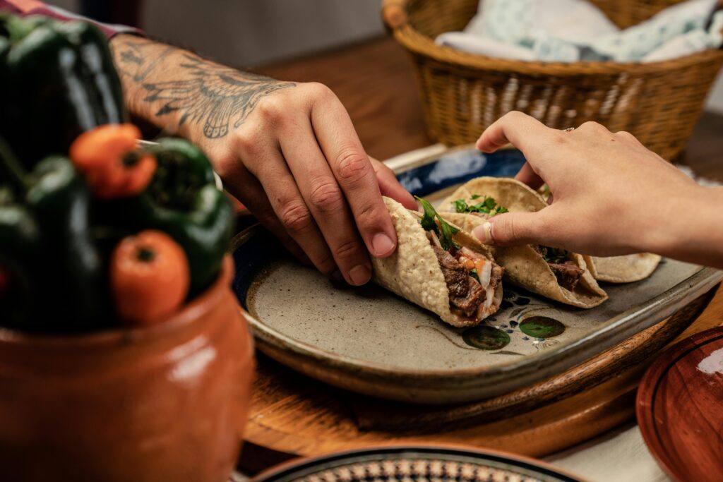 Close-up of hands reaching for tasty Mexican tacos on a ceramic plate, surrounded by fresh ingredients.