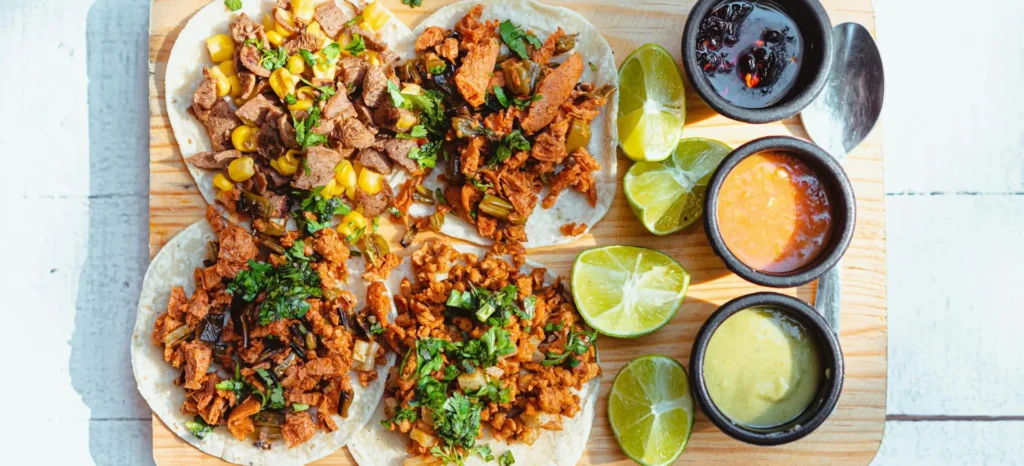 Colorful vegan tacos with lime slices and dipping sauces on a wooden tray, shot in bright light.